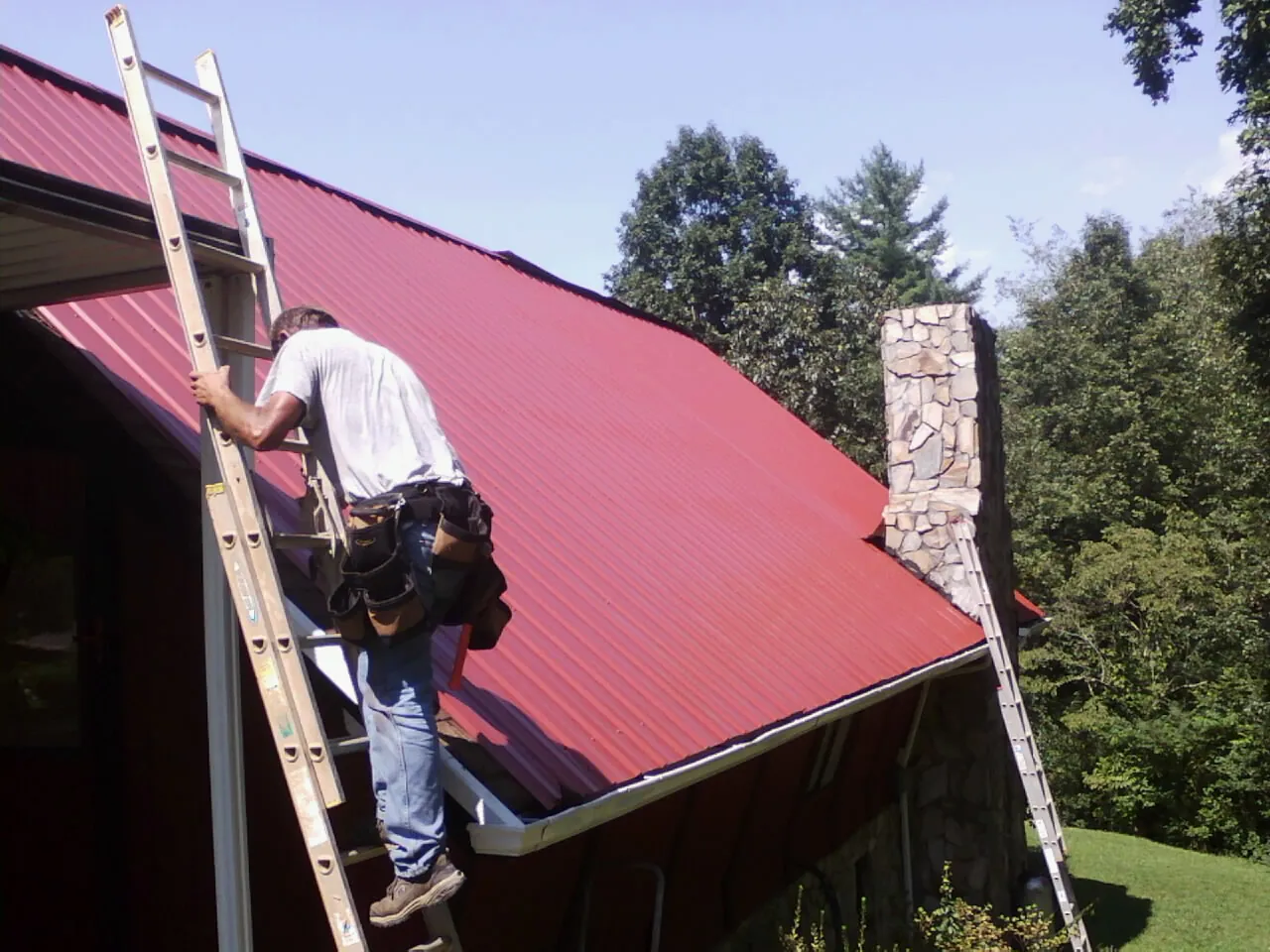Roofer on a ladder beside a red metal roof and stone chimney — Soffit & Fascia Repair by Roofing Brothers in Feura Bush